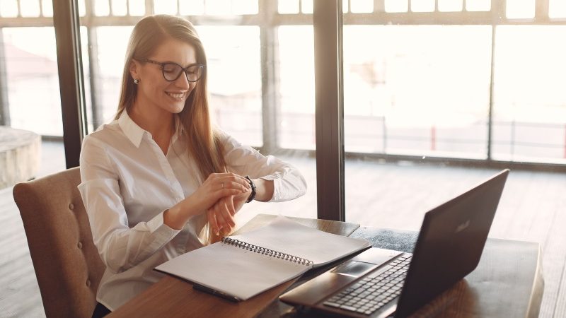 Mulher sentada diante de uma mesa com notebook e livro aberto enquanto consulta o relógio
