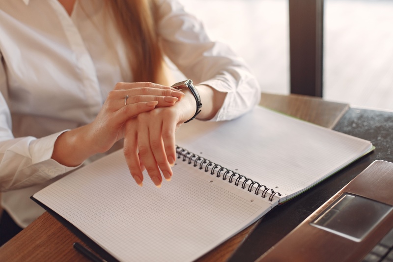 Mulher consultando o relógio diante de uma mesa de trabalho com um livro aberto
