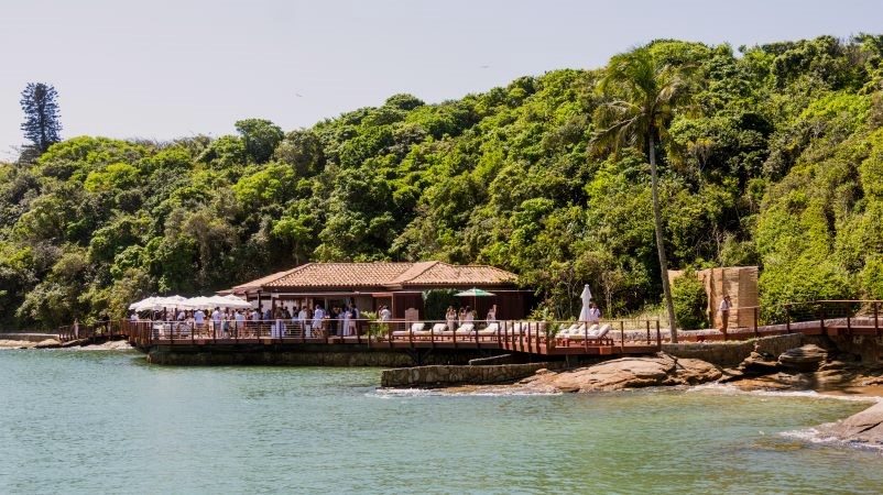 Vista frontal do pier de madeira do Nas Rocas Club, com guarda-sóis e vegetação da Ilha Rasa ao fundo